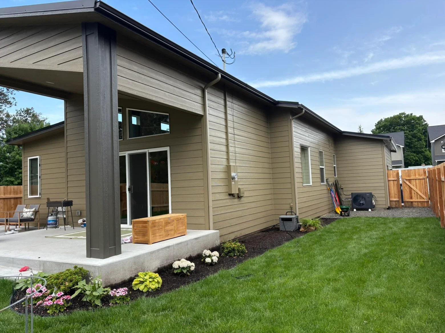 Taupe fiber cement siding with dark trim, covered patio, concrete pad, and landscape beds